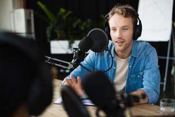 radio host in headphones looking at blurred colleague and gesturing during podcast