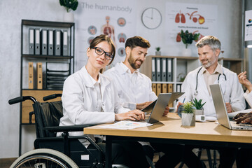 Female doctor using wheelchair typing on laptop and looking at camera while her diverse colleagues chatting near. Group of international physicians having medical training at office room.