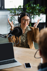 smiling asian announcer in glasses and headphones gesturing while looking at blurred colleague