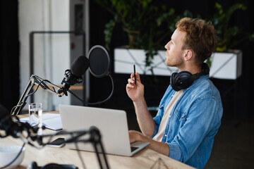 pensive radio host in headphones holding pen near gadgets and microphone in studio