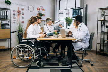 Obraz premium Group of five mixed race medical workers discussing results of x ray scan while sitting together at office room. Qualified doctors and their colleague in wheelchair having working meeting indoors.