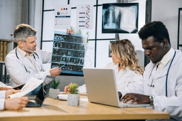 Male and female doctors in white lab coat looking at tomography scan of patient. Group of multiracial colleagues discussing clinical case at office for making diagnosis of severe patient.