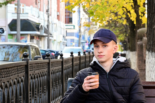 young man in  jacket and baseball cap is sitting on bench and holding paper craft eco cup with drink in his hand. Guy,  student is drinking coffee against backdrop autumn urban landscape. Rest, break