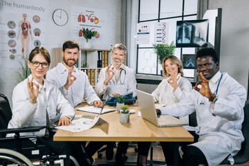 Fototapeta premium Group of five multi ethnic doctors in uniform sitting at table with documents and modern gadgets and looking at camera and showing approval okay gesture. Female young doctor using wheelchair
