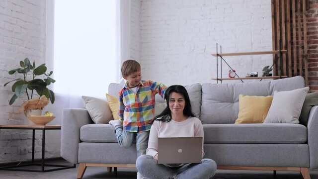 Young Mom sitting on floor using laptop notebook typing message. Happy mother working online at home. Family spending free time at home. Playful kid have fun jumping on leather sofa and laughing.