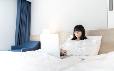 Glasses Asian cute woman works and plays laptop notebook on the bed in Work From Home period.