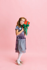 One happy cute caucasian little girl weraing holiday dress standing with huge gift, present box isolated on pink studio background.