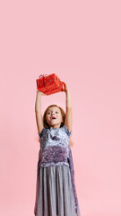 One happy cute caucasian little girl weraing holiday dress standing with huge gift, present box isolated on pink studio background.