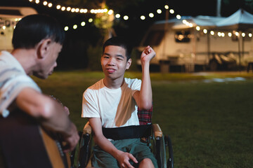 A person with disability in wheelchair singing and father playing guitar happily at the camp night,...