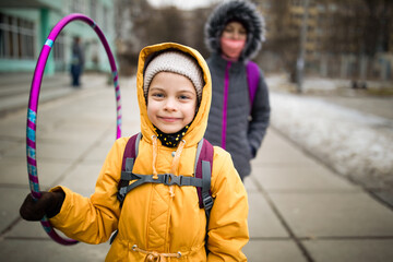 Smiling girl in yellow warm coat, hat and hood holding hoop walking to sports school in winter
