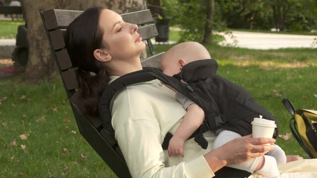 Mother with sleeping baby in infant kid carrier sling resting outdoors in city park, six month old baby boy in baby hipseat. High quality 4k footage