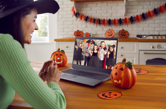 Young teacher happy to see her students at virtual Halloween party for kids. Smiling woman sitting in front of computer at kitchen table decorated with pumpkins and having online party with children
