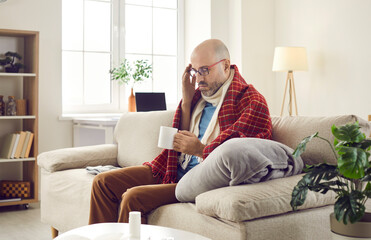 Obraz premium Unhappy tired sick bald mature man who's suffering from common cold or seasonal flu is sitting on sofa at home, wrapped in woolen scarf and plaid, feeling unwell, holding mug and drinking warm tea