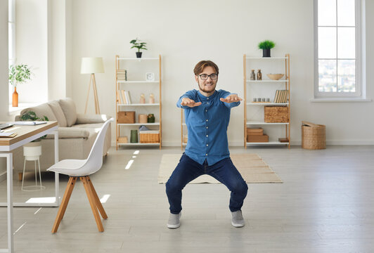 Young active and energetic businessman squatting doing sports training in his home office. Happy man doing squat exercise during working day. Concept of workplace workout for keeping fit and healthy.
