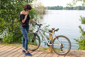 frustrated woman with broken bicycle stands by the river and calls her husband waiting for help.