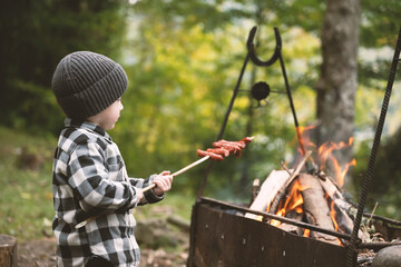 Small kid in a plaid shirt and gray hat fries sausages on campfire on the forest. Childhood with...
