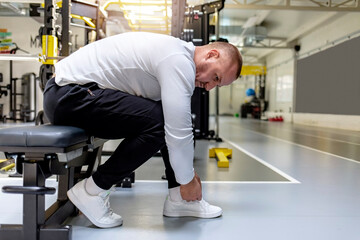 Man tying his shoelaces in a gym. Athletic man tying shoelaces during rest after training in gym while sitting on the bench. Fit young man takes a short break from working out to tie his shoelace.