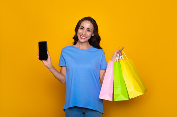 Fototapeta na wymiar Happy smiling young woman in casual clothes with colorful shopping bags is showing smart phone and posing on yellow background