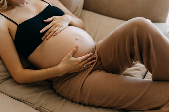 Closeup of a pregnant woman wearing cozy home wear, lying on a sofa at home, holding hands on her belly.