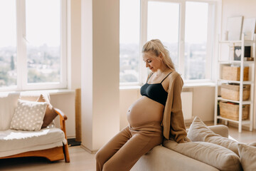 Pregnant woman wearing cozy home wear, sitting on a sofa in light airy interior.