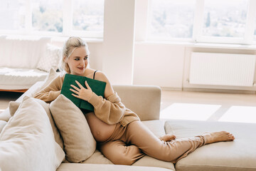 Blonde pregnant woman wearing home wear, sitting on a sofa at home, holding a book.