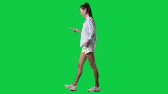 Happy young smart casual summer style woman walking and using cell phone. Side view. Full body on green screen chroma key background