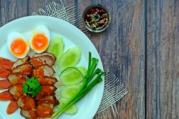 Barbecued red pork, Chinese sausage and boiled egg with rice topped with red sauce served with spring onion and cucumber slices having pickled chilli aside on wooden table.