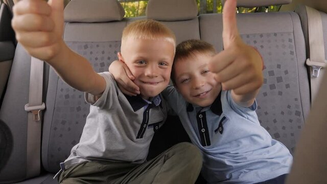 Portrait of two little boys in identical T-shirts in the back seat of a car, they look into the camera and give a thumbs up.