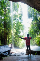 A boy holding a hand stands under a waterfall that falls in front of him.