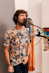 Young caucasian musician singing into microphone while working on songs during a recording studio session at home. Grey background. Millennial singer using headphones, recording his voice.