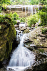 .A small waterfall falls from a cliff and flows through a large rock with turbulent water and green trees.