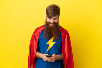Redhead Super Hero man isolated on yellow background sending a message with the mobile