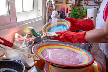Elderly woman washing crockery in front to the window, wearing red gloves. Home kitchen and colorful dishes under flush of water