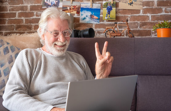Senior attractive man using laptop in video call. Old retired man spending his time by working on laptop at home with camera, decorations, pictures. Brick wall background