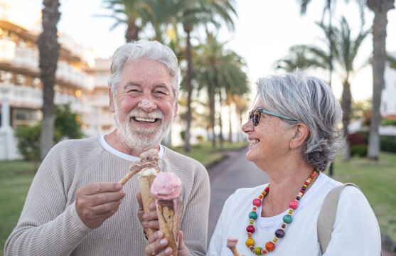 Cheerful senior couple eating ice cream cone in the park. Her wife looks amused at her husband's nose covered with ice cream. Joyful elderly lifestyle concept. Sea and palm trees in the background