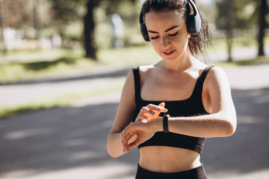Athletic young happy woman runing with headphones in the park, the girl looks at the watch Outdoor workout. Sport and healthy active lifestyle concept. Black clothing for sports
