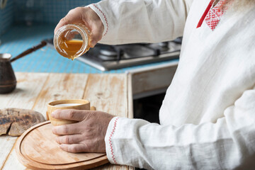 Ethnic linen shirt on the man. A man with a beard in the kitchen pours honey into a cup. Cooking dessert in a rustic kitchen.
