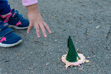 The child's hand reaches for the ice cream that has fallen on the asphalt. Bright ice cream in a green cone fell on the asphalt.
