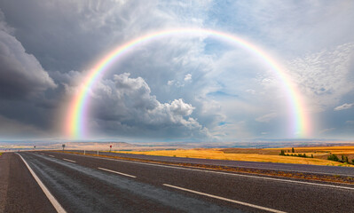 Asphalt road going beyond the horizon at the end of the path amazing rainbow with stormy dark   clouds - Powered by Adobe
