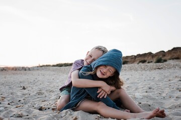 brother and sister sat on sand playfully hugging at the beach