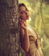Sexy romantic makeup beautiful emotional blond woman posing in fashion gold color dress near the trunk of tree on summer background outside . Closeup