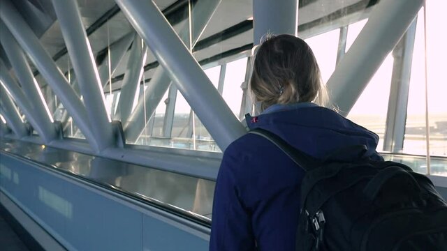 Passenger on airport moving walkway going to flight boarding gate. Young woman traveler with backpack. International terminal interior with sunset light.
