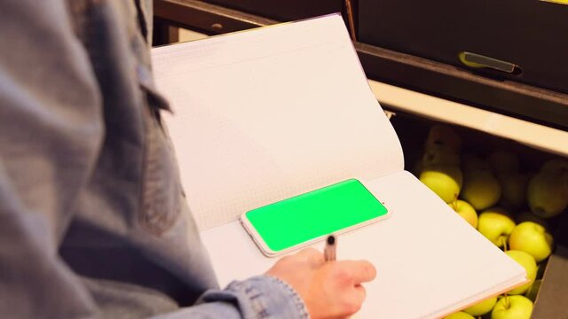 Shop worker making notes with the pen in the journal having the phone on it, while performing inventory of the products.