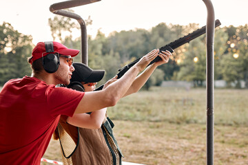 Nice caucasian man in headset is confidently teaching female to use rifle, weapon on outdoor range,...