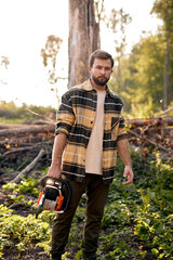 Fototapeta premium Serious young caucasian lumberjack in casual shirt is preparing to cut trees with electric saw, standing in forest alone, posing, at summer evening. outdoor activity. strong man with saw. woodcutter.