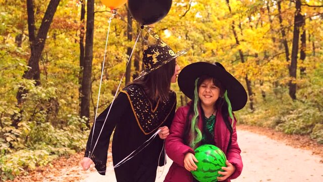 Elder sister saying something funny in ear to her little sister while walking in the autumn forest dressed both in Halloween costumes and holding balloons and watermelon.