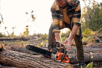 Cropped lamberjack is going to saw branches with chainsaw from felled tree in summer forest. Concept of professional logging, wood working. Young male in casual outfit preparing to chop, cut trees