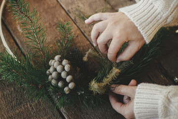 Hands making modern christmas wreath on rustic table with fir branches, brunia herb, round wooden hoop. Atmospheric moody image. Winter holidays preparation close up