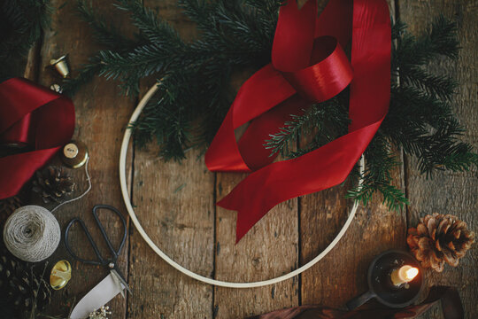 Making stylish modern christmas wreath flat lay. Fir branches, round wooden hoop, red ribbon, candle and pine cones on rustic table. Atmospheric moody image. Winter holidays preparation