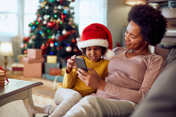 Obraz na płótnie Canvas Happy African American mother and daughter use smart phone while relaxing at home for Christmas.